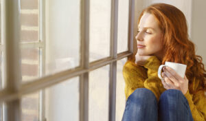 Frau mit roten Haaren in einem gelben Pullover hält eine Tasse und blickt friedlich aus einem sonnenbeschienenen Fenster.