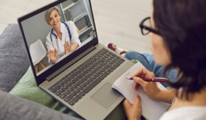 Person, die sich während eines Videoanrufs mit einem Arzt auf einem Laptop Notizen macht.