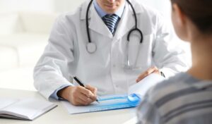 Ein Arzt im weißen Kittel macht sich Notizen, während er in einer Praxis mit einem Patienten spricht.