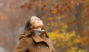Frau im braunen Mantel steht draußen, die Augen geschlossen, lächelt und genießt die Herbstluft mit bunten Blättern im Hintergrund.