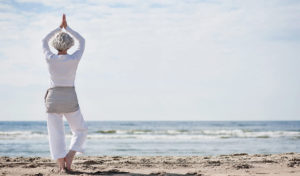 Eine ältere Frau praktiziert Yoga in der Baumstellung an einem Sandstrand mit Blick auf den Ozean unter einem bewölkten Himmel.