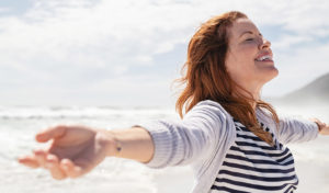 Lächelnde Frau mit ausgestreckten Armen am Strand, die frische Luft und den Sonnenschein genießt.