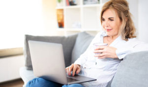 Frau sitzt auf einem Sofa, benutzt einen Laptop, hält eine Tasse in der Hand, schaut auf den Bildschirm und lächelt.