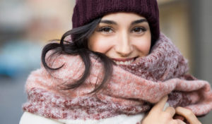 Eine lächelnde Frau trägt im Freien eine kastanienbraune Strickmütze und einen dicken rosa Schal.
