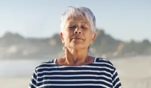 Eine ältere Frau mit kurzen weißen Haaren und gestreiftem Hemd steht mit geschlossenen Augen an einem Strand.