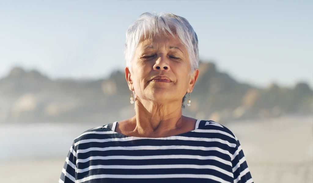 Eine ältere Frau mit kurzen weißen Haaren und gestreiftem Hemd steht mit geschlossenen Augen an einem Strand.