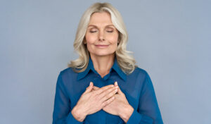 Lächelnde Frau mit geschlossenen Augen hält die Hände auf ihrer Brust vor einem schlichten blauen Hintergrund.