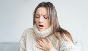 Frau in einem cremefarbenen Pullover, die ihre Brust umklammert und scheinbar Atembeschwerden hat.