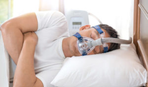 Mann schläft auf der Seite, trägt eine CPAP-Maske, ist an ein Gerät angeschlossen und ruht mit dem Kopf auf einem weißen Kissen.