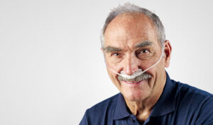 Lächelnder älterer Mann mit einer Nasenkanüle für die Sauerstofftherapie, bekleidet mit einem marineblauen Hemd.