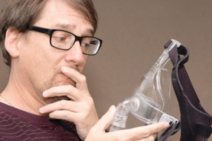 Mann mit Brille betrachtet nachdenklich eine CPAP-Maske in seiner Hand vor einem schlichten Hintergrund.