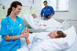 Eine Krankenschwester lächelt und hält die Hand eines Patienten in einem Krankenhauszimmer mit zwei Patienten und einem Arzt.